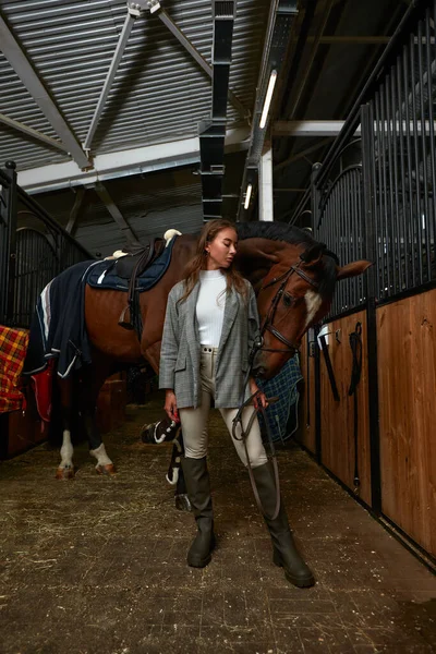 Glimlachende jonge vrouw staan in een stal met een zadel tijdens de voorbereiding van haar kastanje paard voor een ritje — Stockfoto