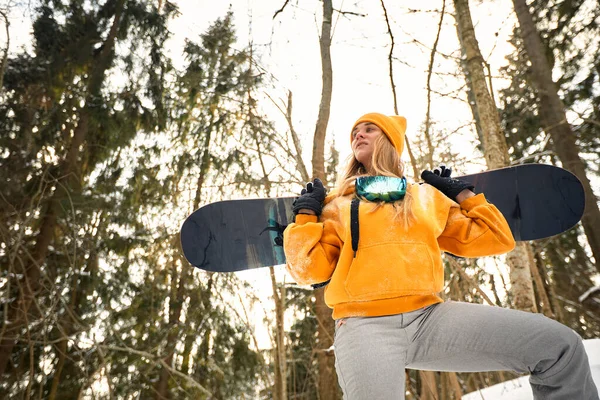 Niña o mujer snowboarder entra en los deportes de invierno en el bosque nevado que se para en la nieve y sostiene una tabla de snowboard —  Fotos de Stock