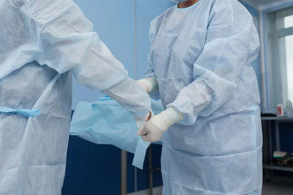 El cirujano y la enfermera se están preparando para la cirugía en el quirófano. Ponte batas, sombreros y máscaras. Instalación de equipos, iluminación, preparación de láminas estériles . — Foto de Stock