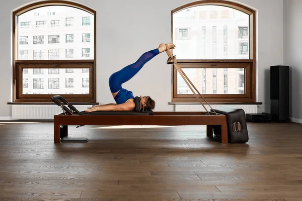 El instructor hace ejercicios sobre el reformador, una hermosa chica entrena en el moderno simulador de reformador para ejercitar los músculos profundos, el equipo de reformador modernizado para Pilates y yoga. — Foto de Stock