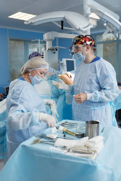 Chirurg a ošetřovatelka se připravují na operaci v operačním sále. Nasaďte si župany, klobouky a masky. Nastavení zařízení, osvětlení, příprava sterilních listů. — Stock fotografie