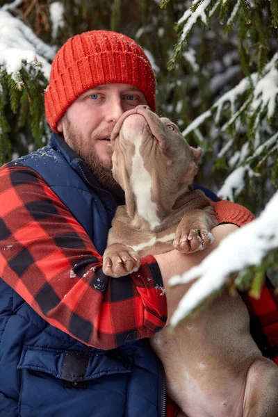 Homem feliz segurando cão adorável em suas mãos na floresta nevada. Garoto sorridente abraçando cachorrinho adorável em madeira de inverno. Amante de animais. Cão - conceito de amigo humano. — Fotografia de Stock