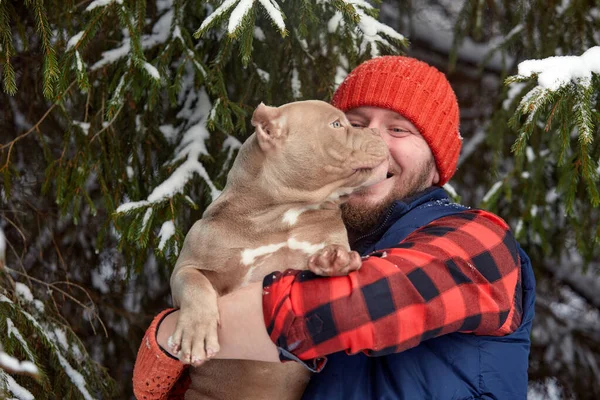 Hombre feliz sosteniendo un perro encantador en sus manos en el bosque nevado. Niño sonriente abrazando adorable cachorro en madera de invierno. Amante de mascotas. Perro - concepto de amigo humano. —  Fotos de Stock