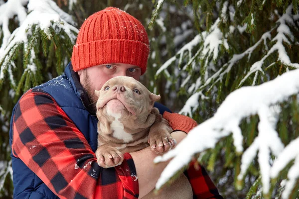 Happy man holding lovely dog in his hands in snowy forest. Smiling boy hugging adorable puppy in winter wood. Pet lover. Dog - human s friend concept. — Fotografia de Stock