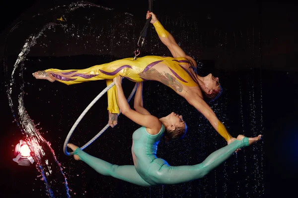 Two Beautiful young weman arobatics in blue and yellow bird color sportswear with halahub in aqua studio. Drops of water spread about her fitness body on the background of water splashes — Stock Photo, Image