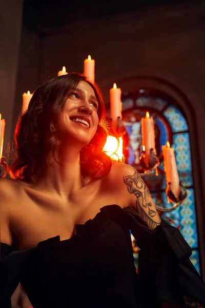 Gothic thema. Mooie jonge dame in het kort zwart jurk zit op stenen sokkel in de buurt van kaarsen op donkere achtergrond. — Stockfoto