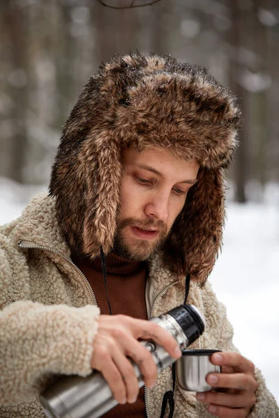 Красивый турист в теплой одежде наливает чай в кружку из термоса в зимнем лесу. Концепция сезона. Селективный фокус . — стоковое фото