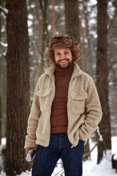 Улыбающийся красивый мужчина в шляпе наслаждается холодной снежной погодой на природе в течение дня в лесу — стоковое фото