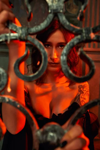 Thème gothique. Belle jeune femme en robe noire courte regarde à travers un treillis sculpté forgé avec des bougies crucifix derrière elle à fond sombre. — Photo