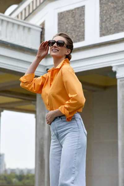Outdoor modeportret van een elegante, luxe vrouw met een zwarte zonnebril, trendy oranje shirt en jeans. Kopieer, lege ruimte voor tekst — Stockfoto