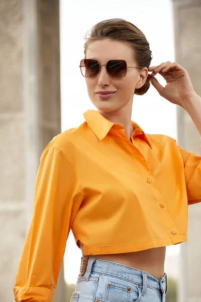 Ritratto di moda outdoor di donna elegante e di lusso con occhiali da sole neri, camicia arancione alla moda e jeans. Copia, spazio vuoto per il testo — Foto Stock
