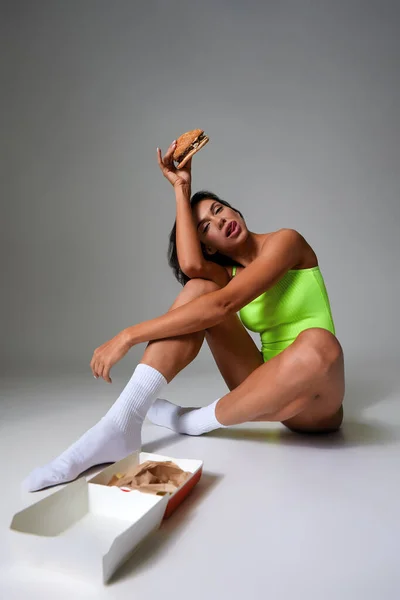 性感运动的女人，在灰色的背景上吃汉堡包 — 图库照片