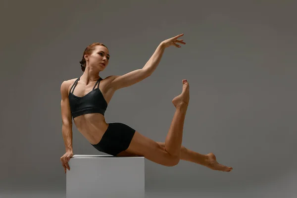 Beautiful athletic woman gymnast in sportswear performs gymnastics fitness exercises on a gray background. Sports motivation, stretching.