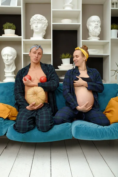 Funny pregnant girl with her husband play. Pregnant woman holds her belly and her husband holds a pumpkin instead of his belly and tomatoes at his chest.