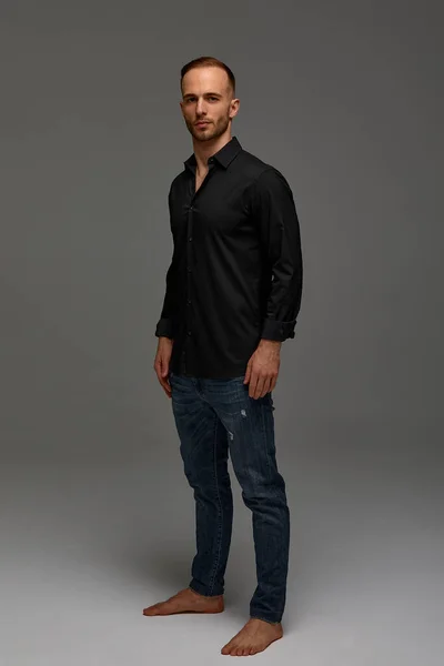 An attractive white man in jeans and black shirts tands in full growth against a gray background. Copy space.