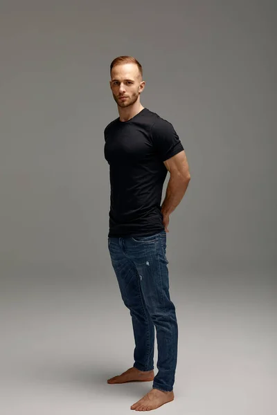 An attractive white man in jeans and black shirts tands in full growth against a gray background. Copy space.