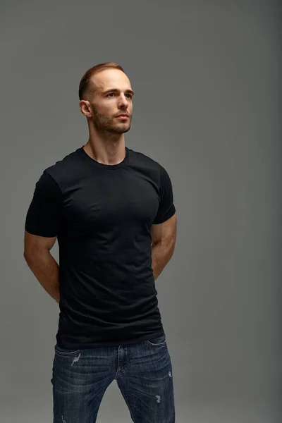An attractive white man in jeans and black shirts tands in full growth against a gray background. Copy space.