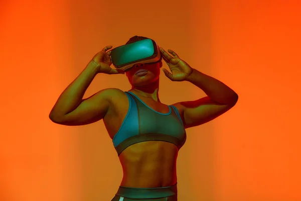 Modern african black woman with short hiar in sport top wearing glasses of virtual reality on yellow neon light background.