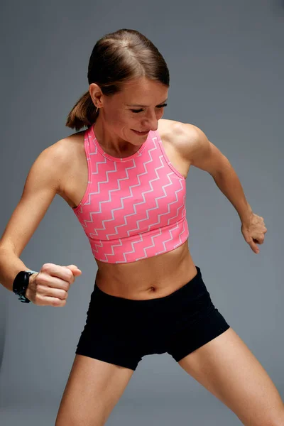 Determined muscular female athlete workout, raising leg and doing stretching exercises. Sport woman in sportswear training indoors, doing fitness aerobics, gray background