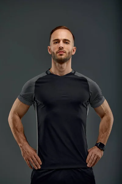 Half length portrait of young sporty guy in t-shirt and shorts standing on gray studio background. Serious millennial sportsman looking at camera. Healthy lifestyle and sports concept.