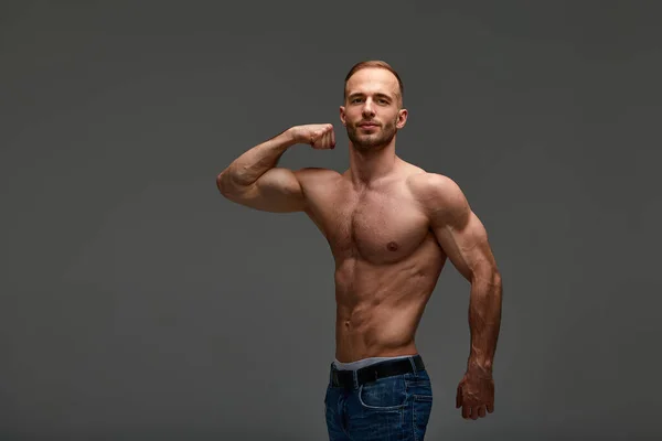 Portrait of a handsome young guy model with hairstyle in a shirtless with a muscular healthy body, showing off his biceps muscles on a gray background