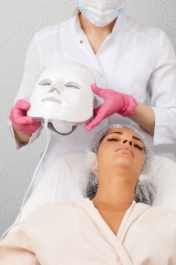 LED light anti-aging mask for facial skin care in a spa slow motion. A woman lies on a couch in a special mask. Modern technologies of beauty and health clipart