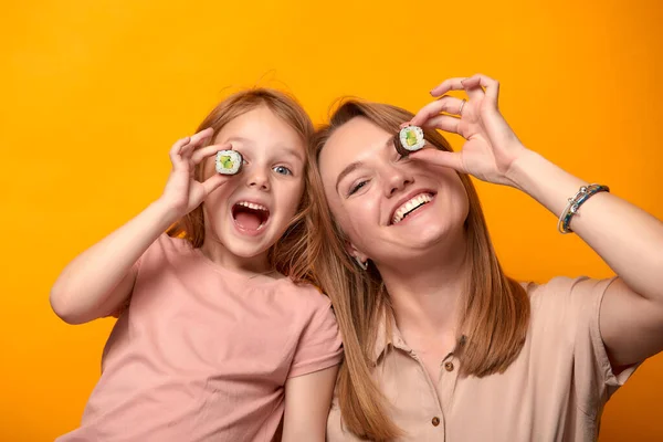 Funny mom with daughter holding sushi rolls in front of eyes on yellow background.