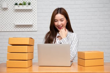 Genç Asyalı kadın, evdeki oturma odasındaki ahşap masada, paket kutusu ve bilgisayar dizüstü bilgisayarıyla kendi başına küçük bir iş kuruyor. Online pazarlama kutu teslimatı kavramı.