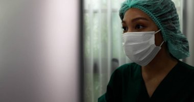 Yüz maskesi takan Asyalı bir hemşirenin portresi doktor odasında oturuyor ve bilgisayarındaki randevularını kontrol ediyor.