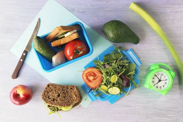 Lunchbox with cereal bread sandwich with cheese, organic vegetables and micro greens, apple, stationery and alarm clock on wooden table, top view