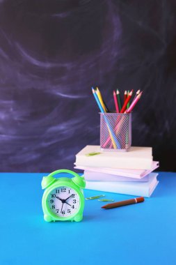 Arka planda bir yığın kitap, kalem ve karatahta, masanın üstünde, okula, eğitime, çalışmaya geri dönen çalar saat.