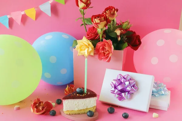 A piece of berry cake with a burning candle, a bouquet of fresh roses, gifts, balloons and festive flags on a bright background, Happy birthday, congratulations concept, postcard 