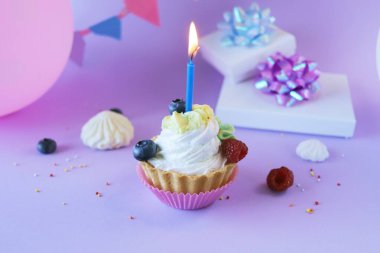 Dessert with white cream decorated with fresh berries and burning candles, lilac background, balloons and gifts with shiny ribbons, happy birthday, holiday, congratulation, postcard