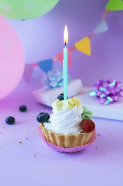 Dessert with white cream decorated with fresh berries and burning candles, lilac background, balloons and gifts with shiny ribbons, happy birthday, holiday, congratulation, postcard