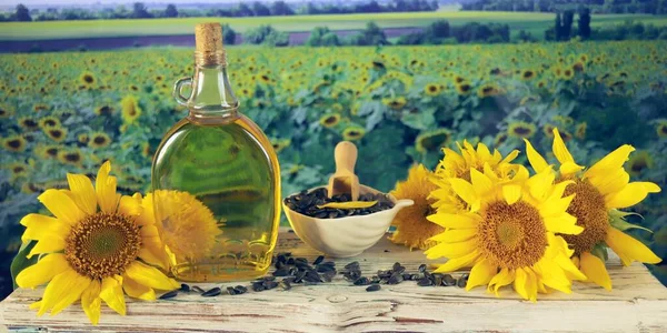 A bottle of oil, fresh sunflower flowers and seeds on the table, against the backdrop of a landscape with a flowering field, organic products, harvest, farm, sale, agriculture