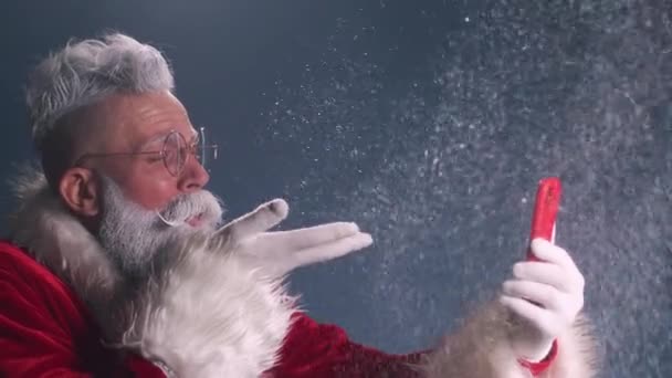 Nowoczesny Wesoły Święty Mikołaj trzyma telefon, wieje śnieg i wskazuje na ekran. — Wideo stockowe