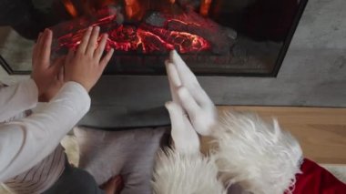 Kırmızı kostümlü Noel Baba küçük çocuklarla şöminede ısınıyor..