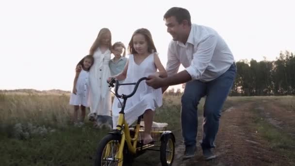 Father with family teaches daughter to ride bike on summer field at sunset. — Stockvideo