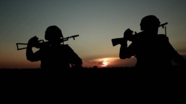 Silhouettes of two soldiers with automatic rifle walking across field at sunset.