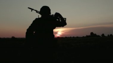 Silhouette of sniper soldier walking at sunset across field with weapon rifle.