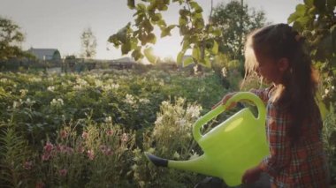 Portrait of happy little girl pouring flowers and fresh fruit, vegetables holding watering can for plant growth in sunshine outdoors. Summer agricultural household activities, farm child work game