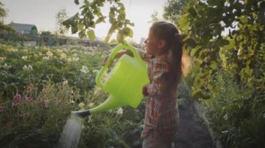Happy smiling child girl helper pouring fresh greenery, vegetables and flowers holding watering can in sun flare outdoors. Garden bed care for plant growth, rural summer work, farm nature activity