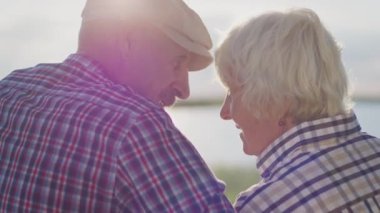 Happy affectionate elderly grandparents together hugging sitting at lake beach date at sunset close up outdoors. Carefree caressing love, calm tenderness, sensual cuddle and support of two old spouses
