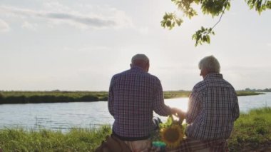 Back view at happy old grandparents holding hands sitting on lake beach in sunshine outdoors. Romantic family relax at nature, calm date walk together, loving support and trust, life harmony feelings