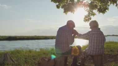 Happy affectionate elderly grandfather kiss grandmothers hand sitting at lake beach in sunshine outdoors. Romantic tenderness, gentle marriage harmony, loving caring feelings, carefree date at nature