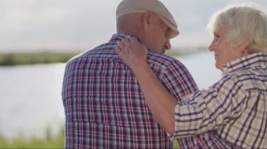 Happy smiling old couple hugging sitting on lake beach in sunshine close up outdoor. Loving harmony, marriage idyllic, romantic relationships, care and support, tender embraces of married grandparents