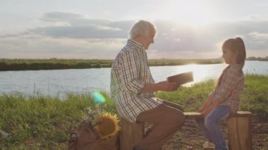 Happy smiling family spend time together outdoor : old caring grandmother read book story granddaughter at sunset. Relationship harmony of grandparents and children, imagination fantasy, peaceful life