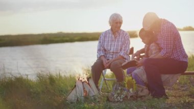 Happy old caring grandparents read interesting book story lovely child granddaughter sitting at lake beach near bonfire at sunset outdoors. Peaceful family rest at nature, child adventure imagination