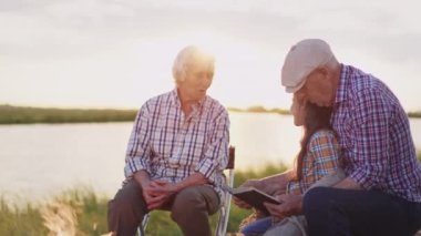 Old smiling grandparents embrace, read inspire book story little joyful granddaughter sitting on pond shore at sunset outdoors close up. Summer family walk : funny adventure hike, calm relax together