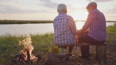 Old affectionate spouses together hug sitting on pond shore near bonfire, contemplating nature beauty at sunset outdoor. Real love, care and support, calm family relax, happy nostalgia at evening walk